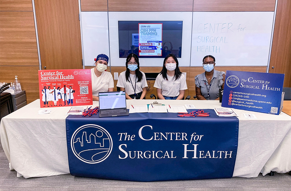 Four young people wearing masks sit at a table with a Center for Surgical Health banner and signs. 